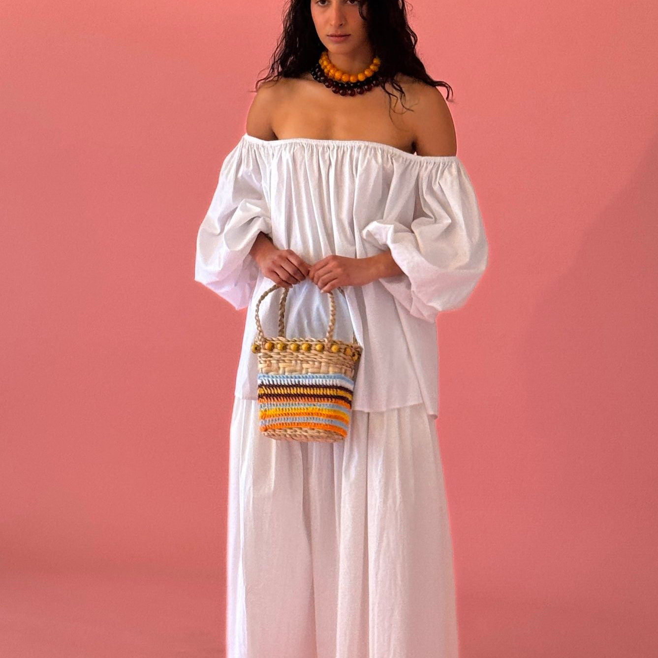 Woman in a white off-shoulder dress holding a colorful woven handbag - Isla Basket bag in yellow against a pink background